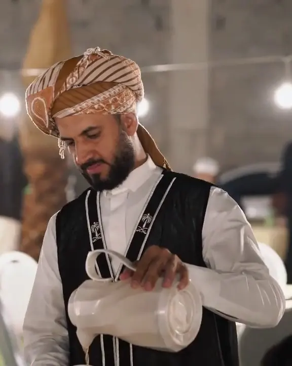 A host in traditional dress pours Saudi coffee under string lights